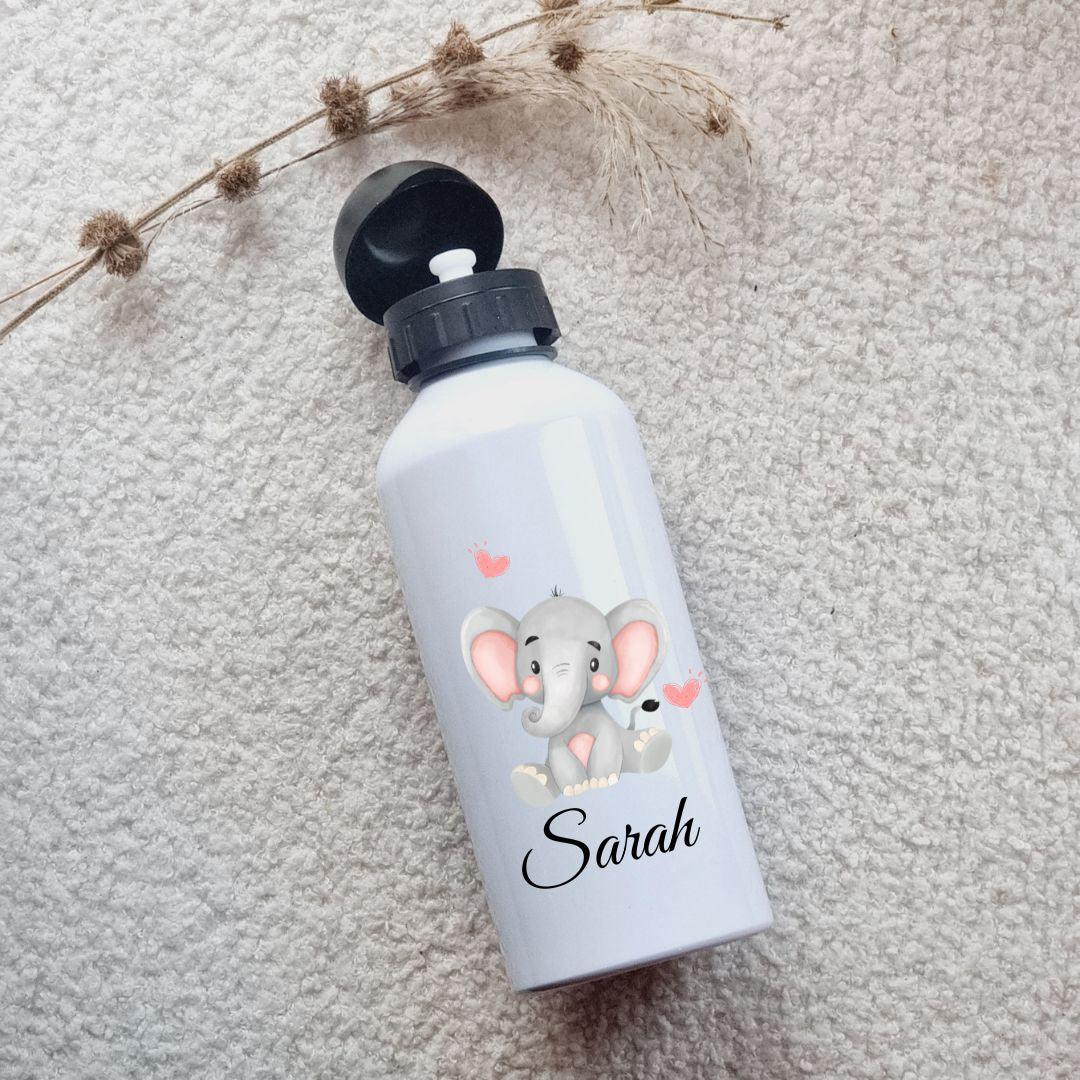 Kit de rentrée scolaire comprenant une boîte à lunch et une gourde assorties, personnalisées avec un prénom et des motifs colorés, posées sur une table en bois avec un fond lumineux.