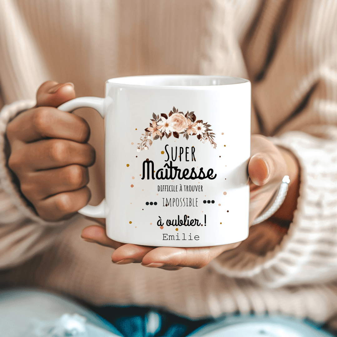  Mug blanc avec l’inscription "Super Maîtresse – Difficile à trouver, impossible à oublier" posé sur un bureau avec des fournitures scolaires.